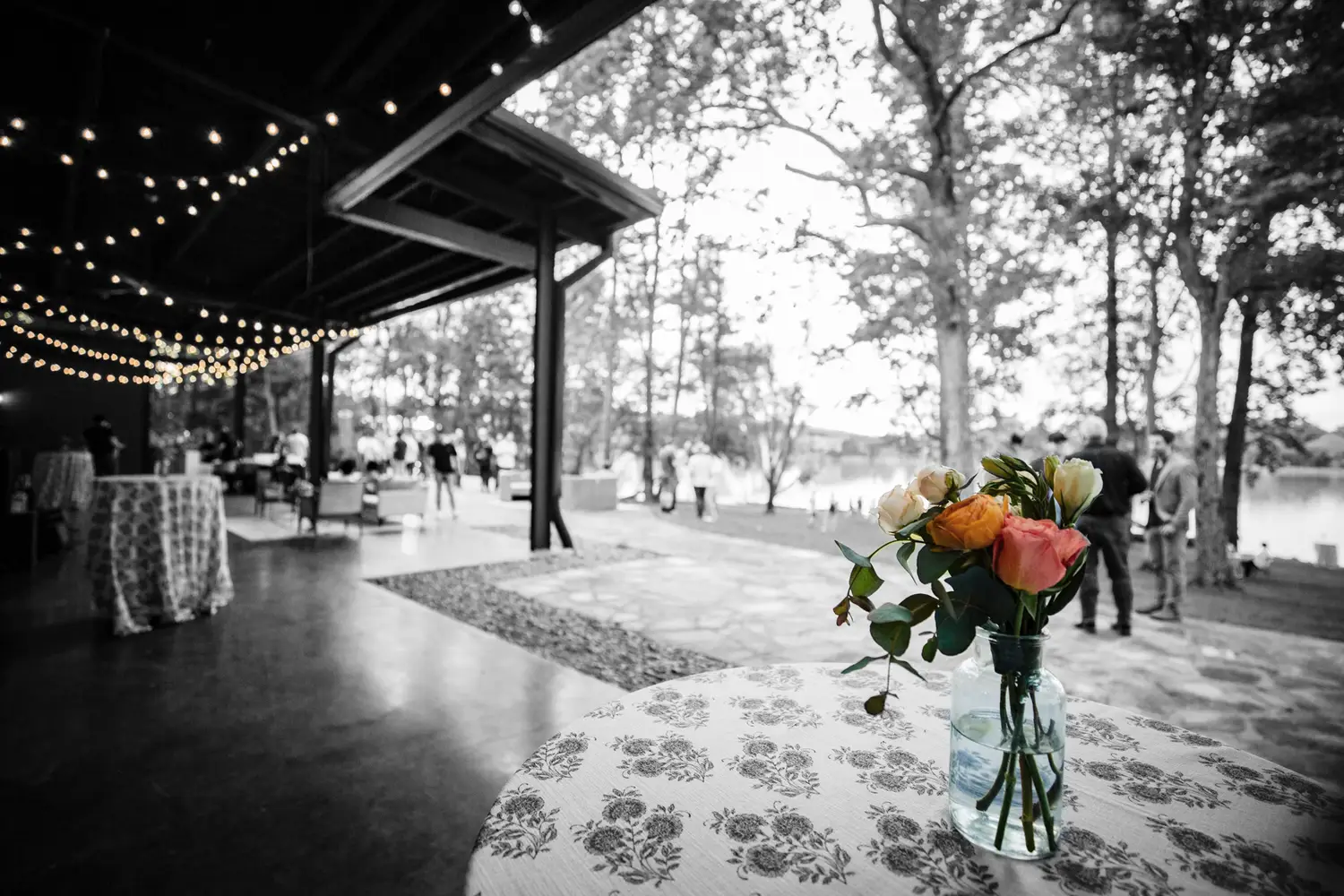 An elegant outdoor reception setting with flowers, lights, and guests near the water.